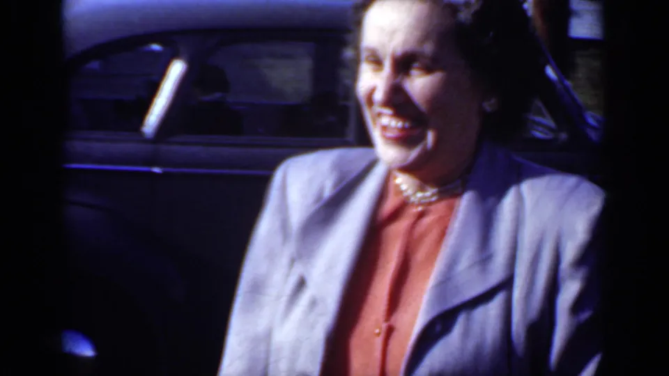 A close frame shows the smiling woman in a grey suit and pearl necklace beside the dark car, with strong sunlight and soft 8mm color.