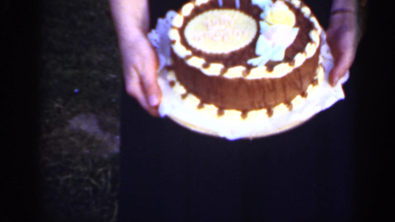 Hands hold a round decorated cake with candles against a dark dress.