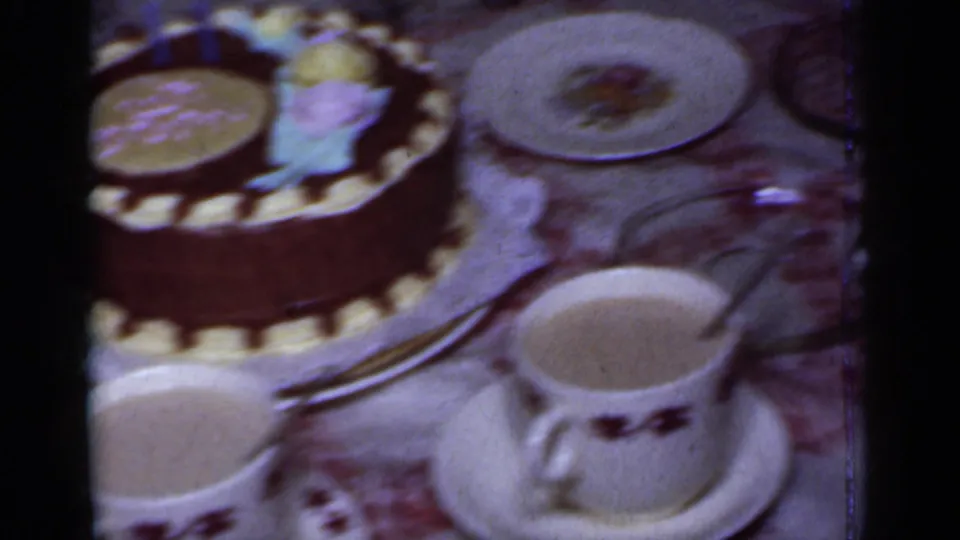 The birthday cake appears beside teacups, saucers, plates, and a patterned table covering in the New York home-movie footage.