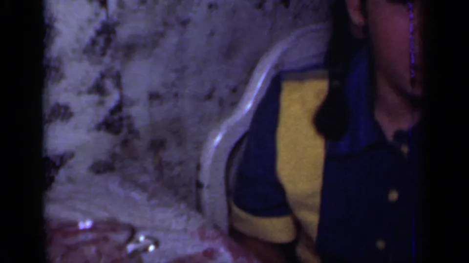 A seated person is partly visible beside the birthday table, wearing blue and yellow clothing in the 1950s New York home-movie clip.