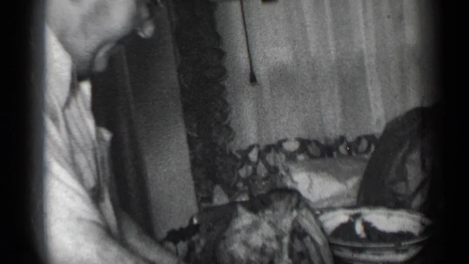 In 1950s New York home-movie footage, a man in glasses bends over a table to cut meat near patterned curtains.