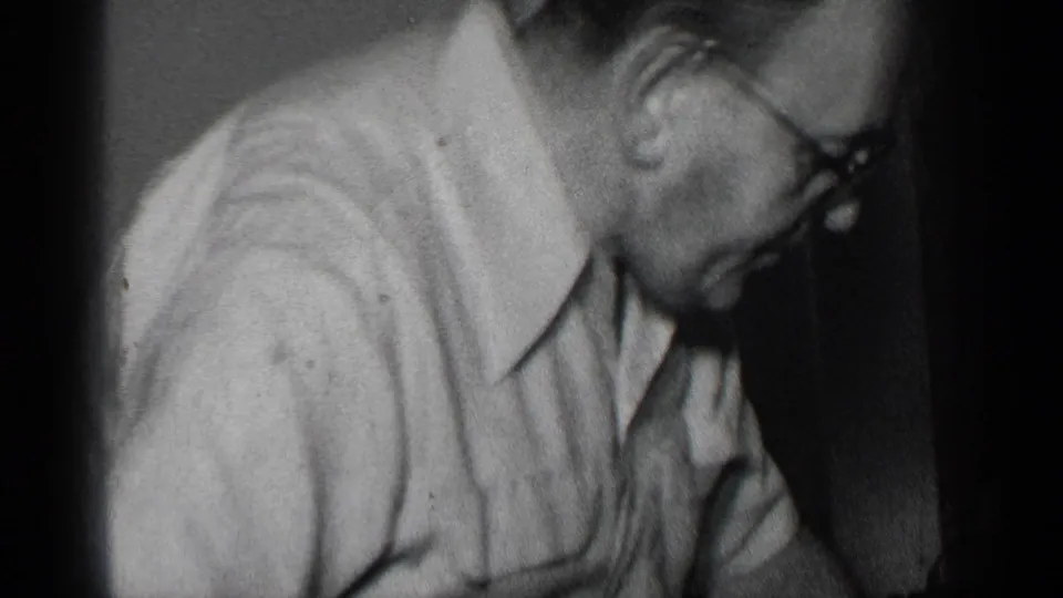 A man wearing glasses leans down beside the table as he works on the turkey in black-and-white 8mm footage.