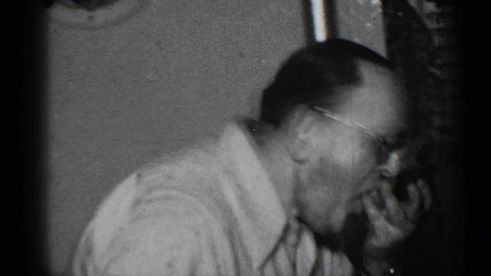 A man in glasses leans forward to taste food in a private interior, with a lamp and wall visible behind him.