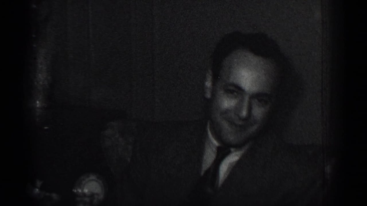 Man in a suit and tie seated indoors, smiling faintly in a dark 8mm frame.