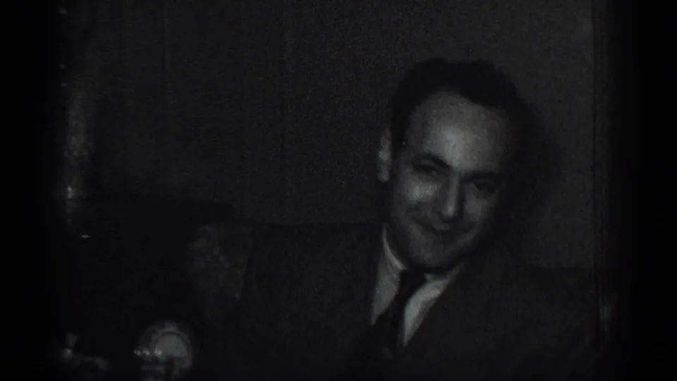 In 1950 New York home-movie footage, a seated man in a suit and tie faces the camera in a dim indoor setting.