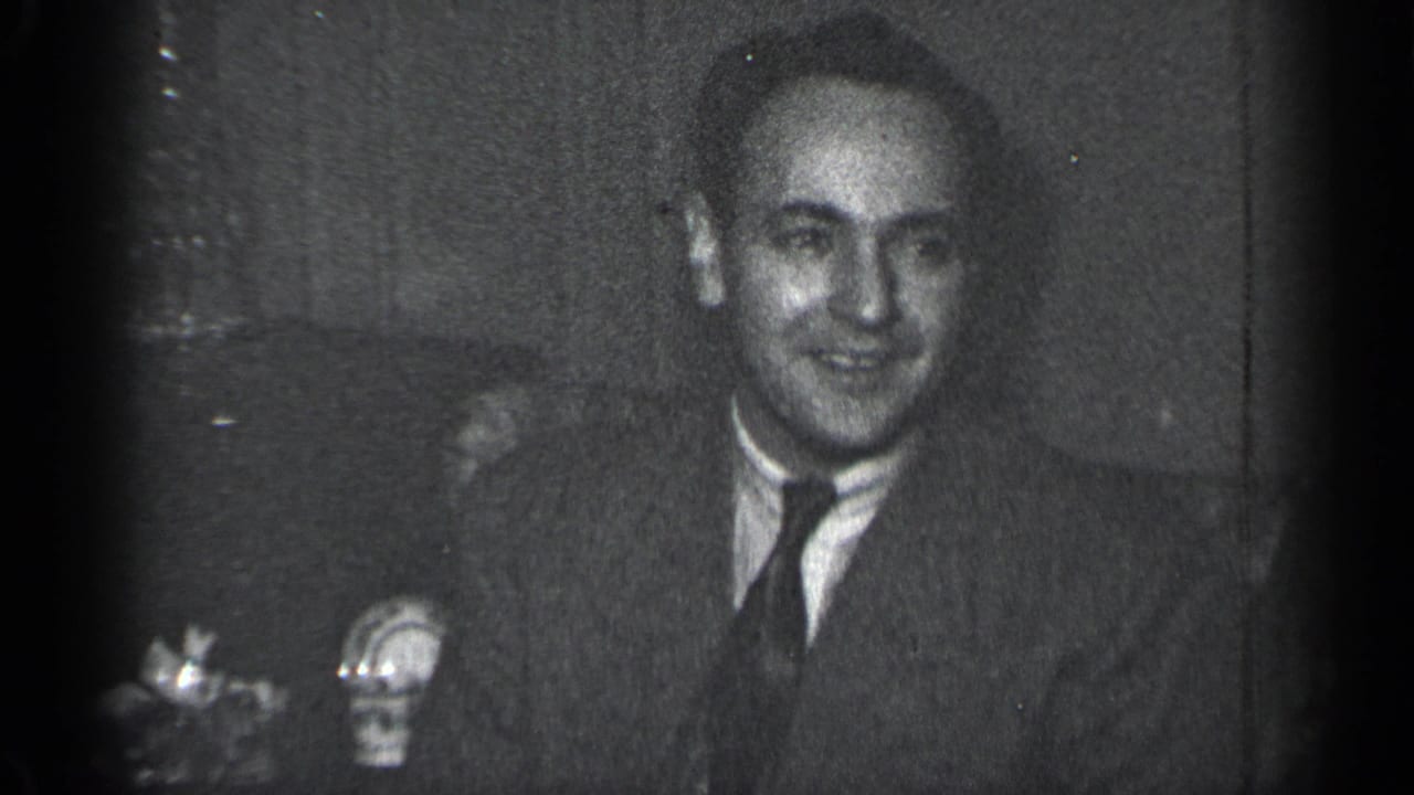 Smiling man in a suit seated indoors beside small tabletop objects.