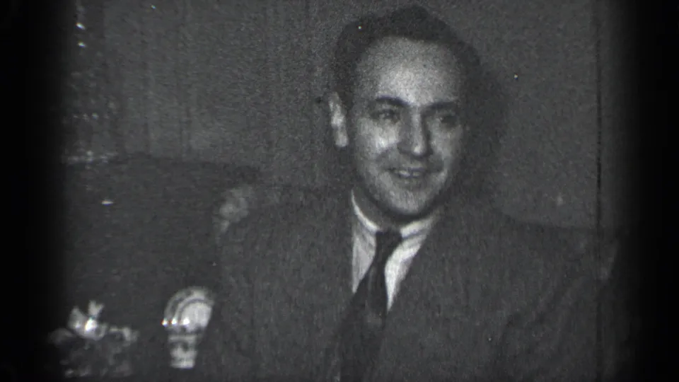 A suited adult man smiles toward the camera in black-and-white 8mm footage from a New York indoor setting.