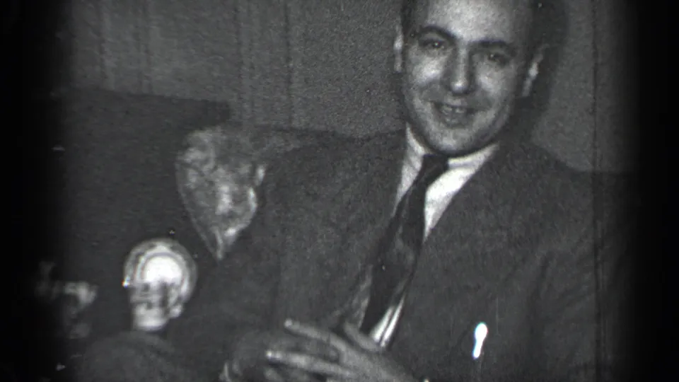 A seated suited man smiles in a private indoor space, with tabletop objects and a textured background visible.