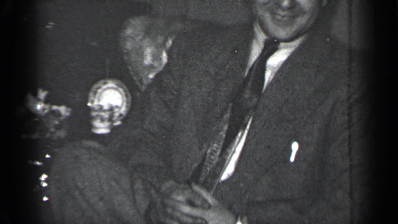 Two suited men seated indoors, one laughing while holding a small object.