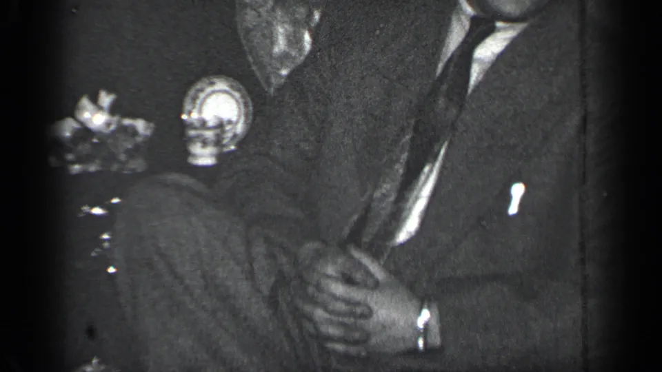 Close 1950 New York home-movie view of suited men seated indoors, with hands and a small object visible near the lap.