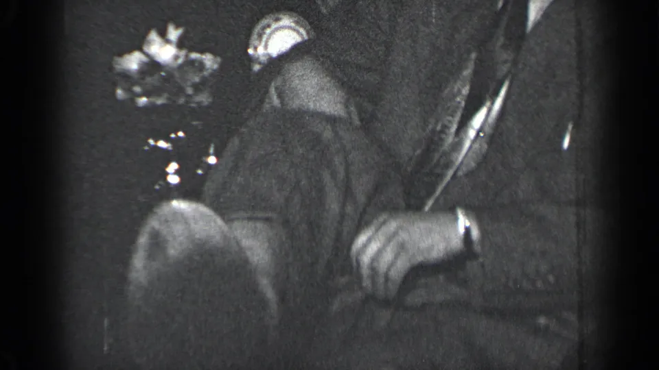 Grainy 8mm frame from 1950 New York showing a suited man seated indoors, head lowered, hands near a small object.