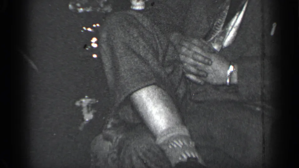 1950 New York home-movie frame of a suited man seated indoors, hands clasped, with another person partly visible nearby.