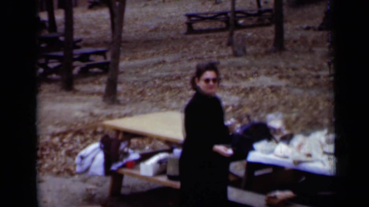 Woman in a black coat and sunglasses standing near picnic tables in a wooded park.