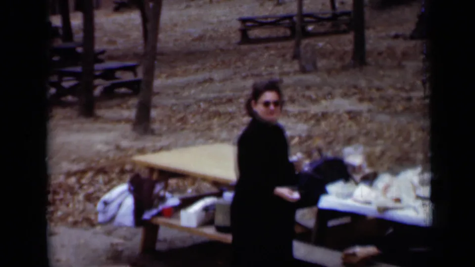 In 1950 New York home-movie footage, a woman in a black coat and sunglasses stands by picnic tables with food in a wooded park.