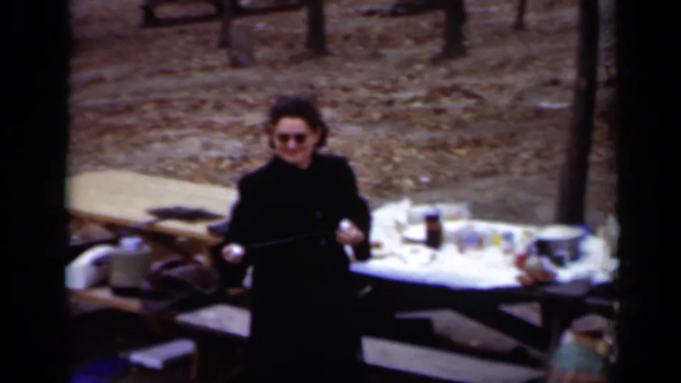 The woman stands near picnic tables in the wooded New York park, holding a long object while food and containers sit on the tables.