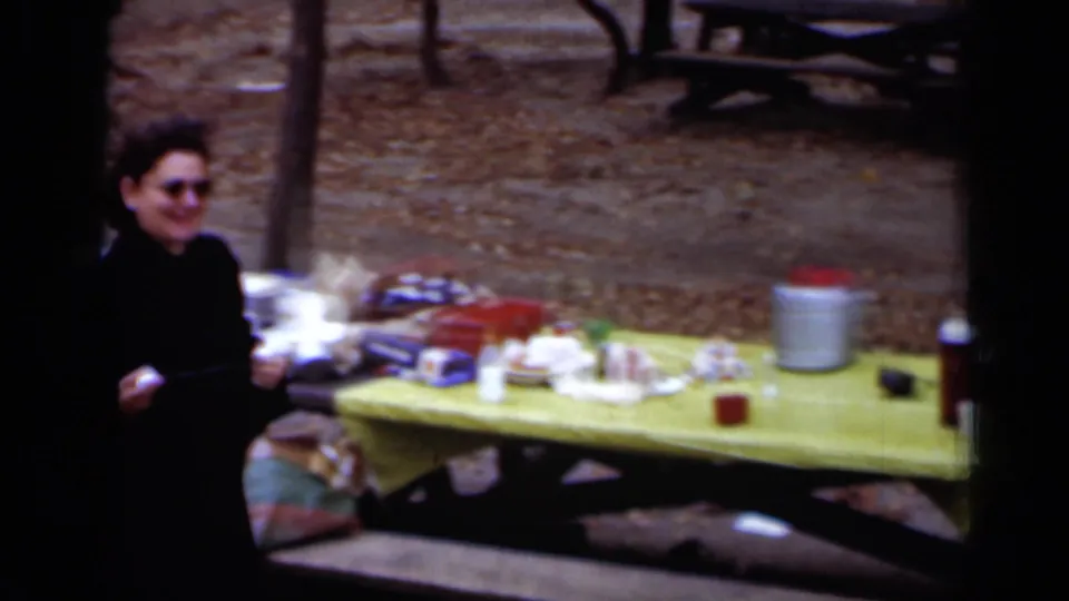 A woman in dark clothing stands at the edge of a picnic table covered with food, a jar and containers in the wooded park setting.