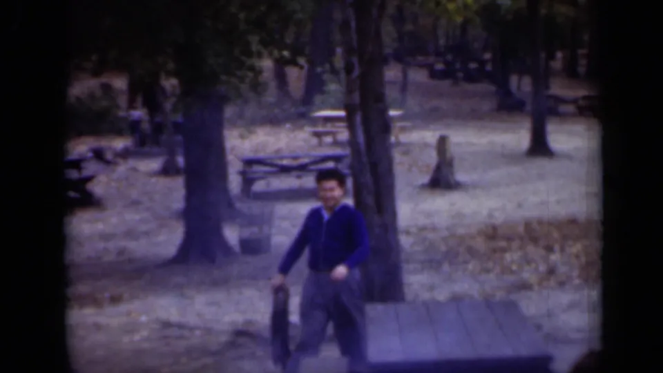 A man in a blue sweater walks through the same New York park picnic area, with trees, benches and picnic tables visible behind him.