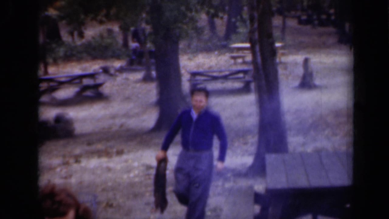 Man in a blue cardigan walks through a wooded park with picnic tables behind him.