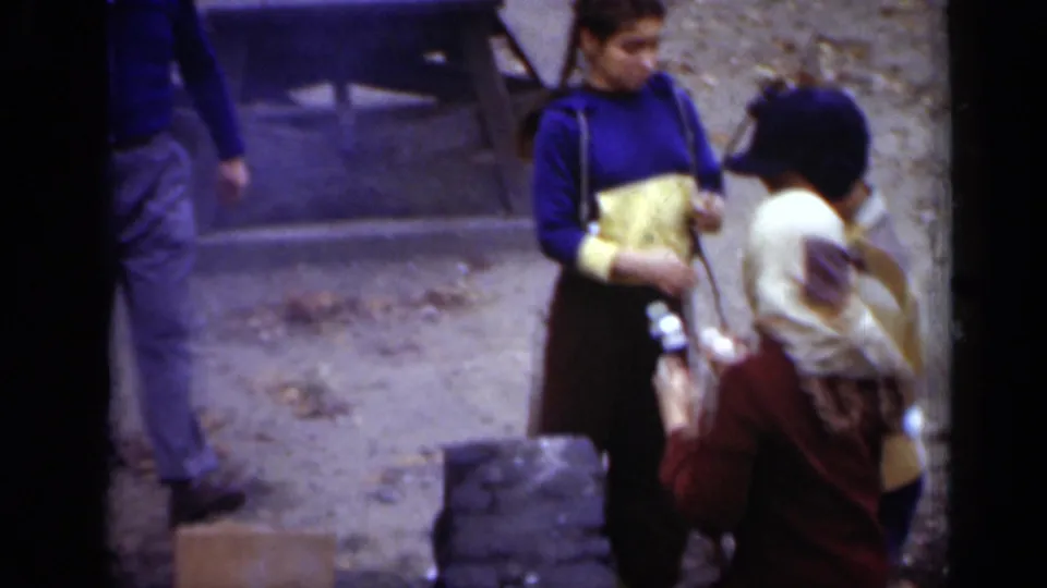 A small group stands near a stone fire pit in the wooded New York park, with one woman holding a bag or folded item.