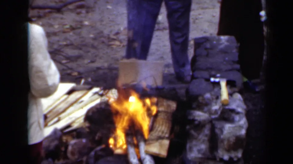 A fire burns in a stone fire pit as people stand close by in the wooded New York park setting.