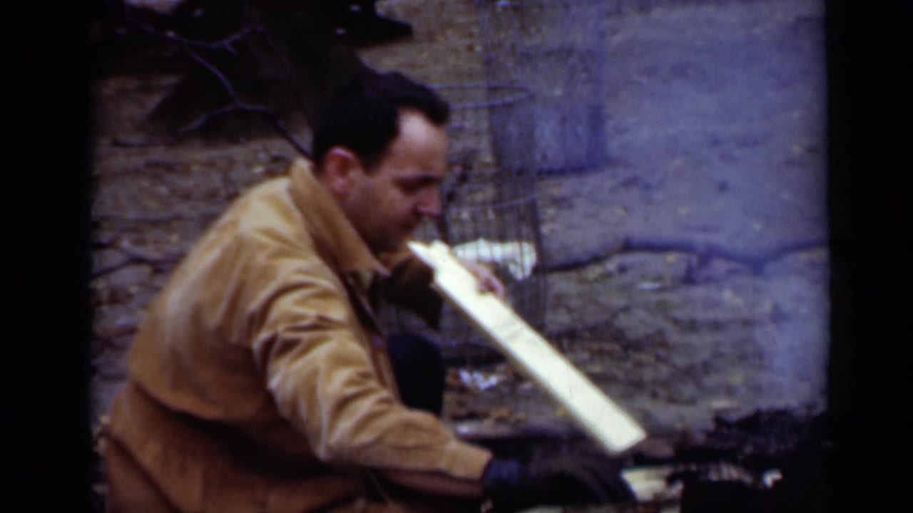 Man in a tan jacket crouches beside split wood near a small outdoor fire.