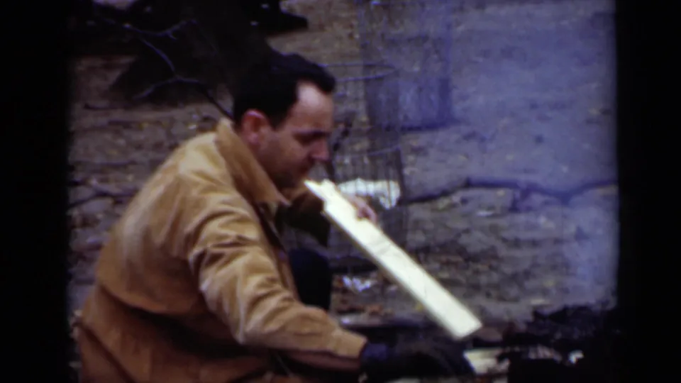 In 1950 New York home-movie footage, a man in a tan jacket and dark gloves crouches near split wood by a small campfire.