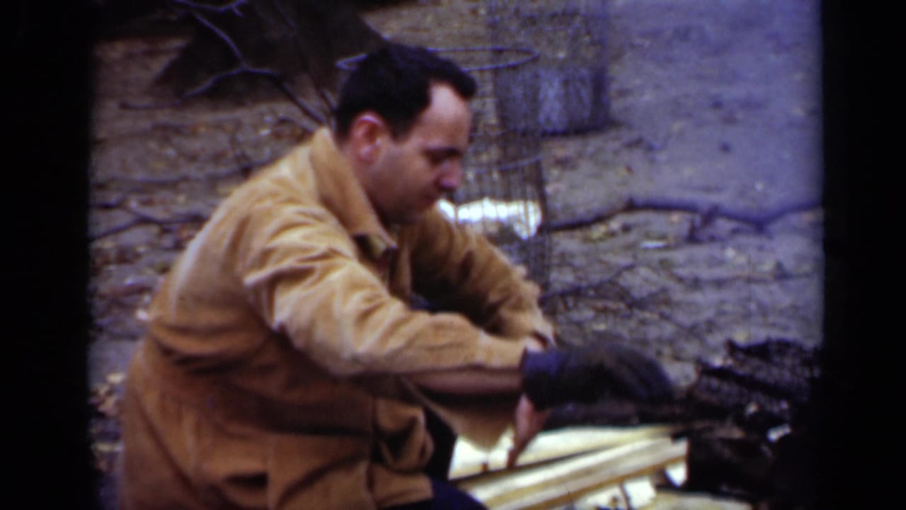 Man reaches toward split wood while crouched beside an outdoor campfire area.