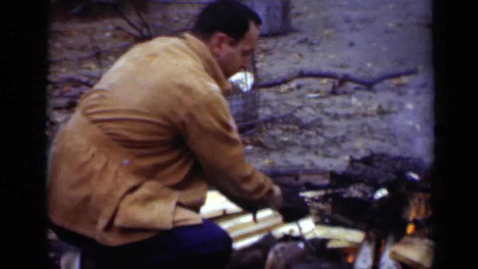 A man in a tan jacket bends over the campfire, moving split wood beside small visible flames in the 1950 home-movie frame.