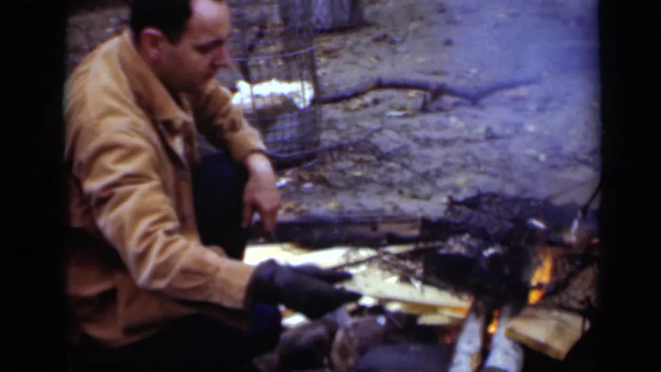 The man crouches at the campfire in New York, extending a gloved hand toward charred wood and split lumber.