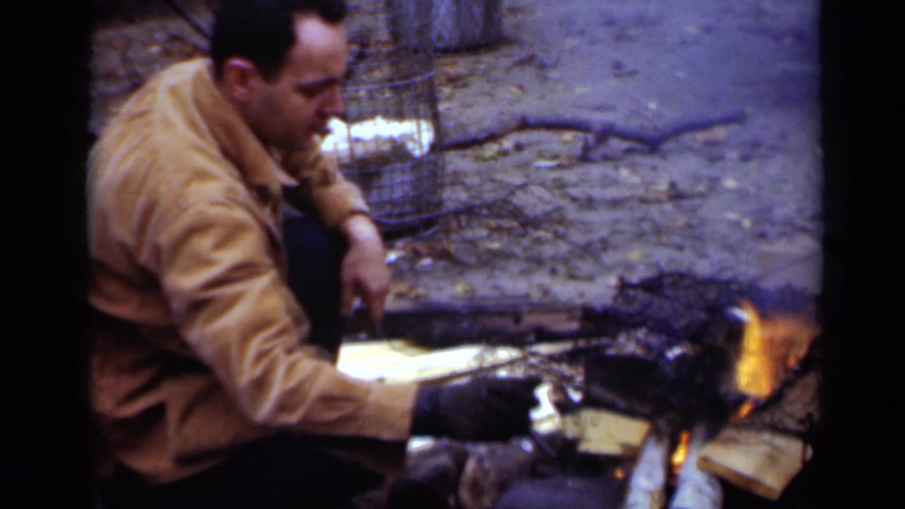 Man in a tan jacket crouches beside a small outdoor fire and charred wood.