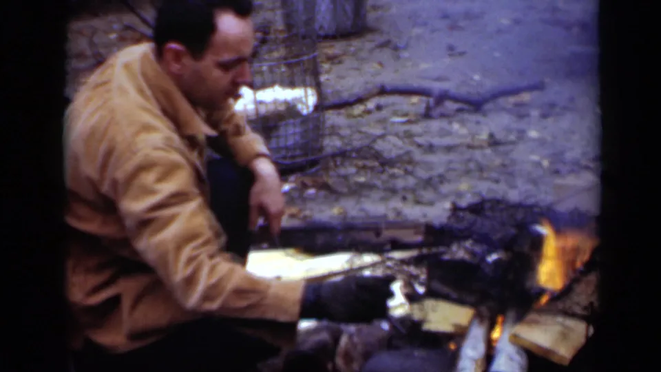 1950 New York 8mm frame of an adult man in a tan jacket and dark gloves crouching by a small fire with wood and debris nearby.