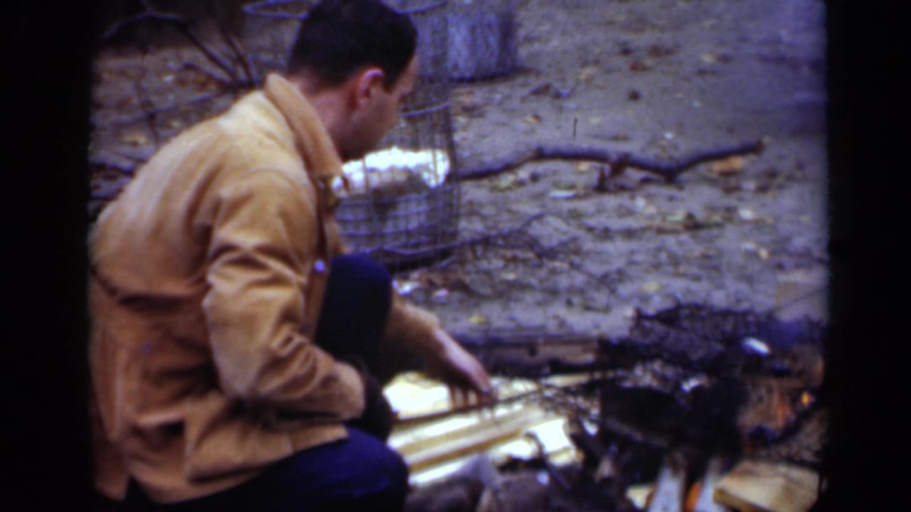1950 New York home-movie frame showing a man in profile crouched near a small fire, with split boards, charred debris, and a wire cage behind him.
