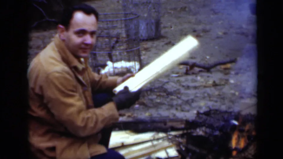 1950 New York 8mm frame of the man turning toward the camera with a pale board in his gloved hands near a small outdoor fire.
