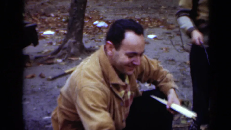 1950 New York home-movie frame with the smiling man crouched near a tree, holding wood beside the small fire area as another partial figure stands nearby.