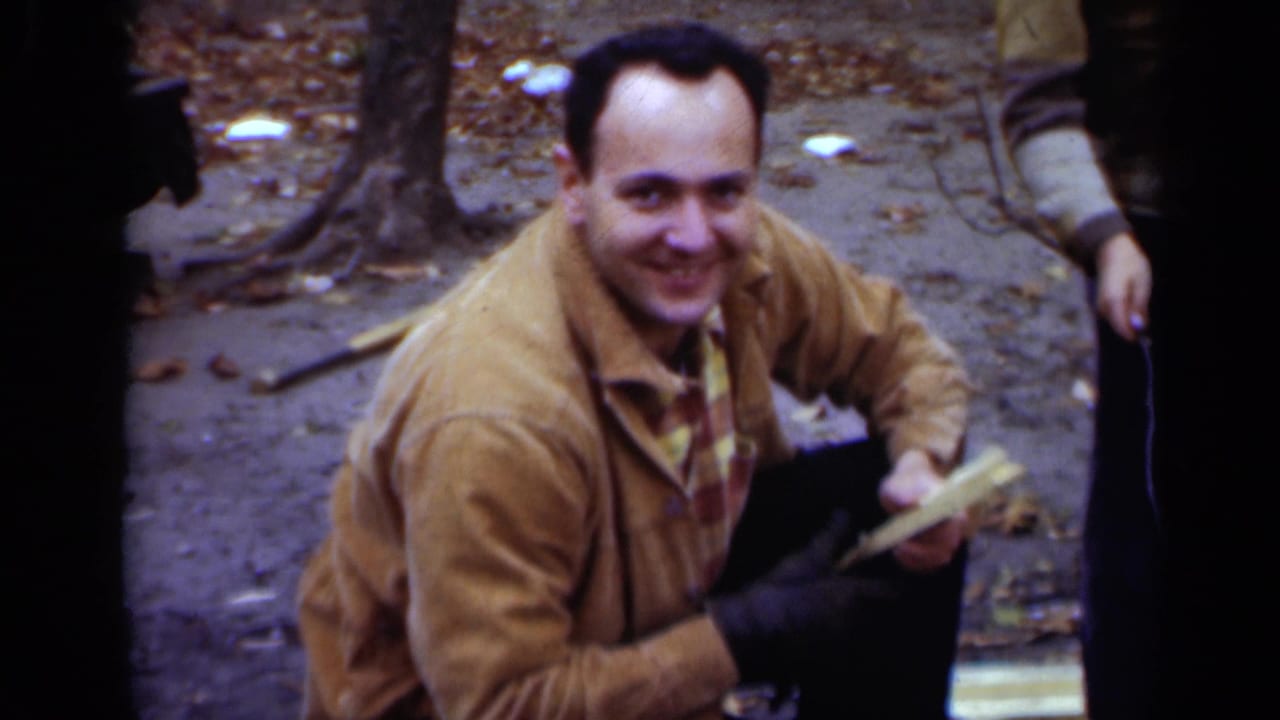 Man in a tan jacket crouches in woods holding a roasting stick near other people.
