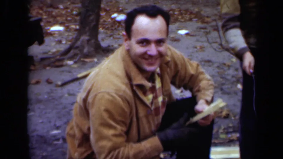 In supported 1950 New York footage, a smiling man in a tan jacket crouches outdoors with a roasting stick while others stand nearby.