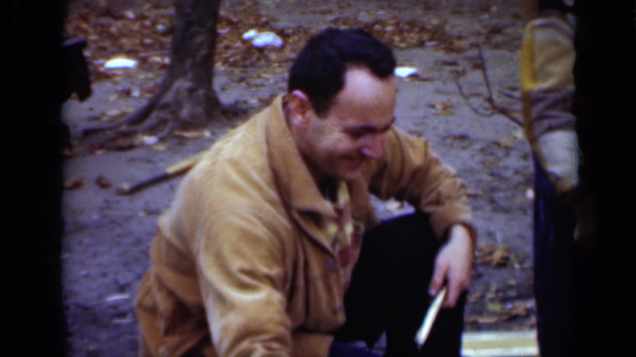 Man crouches beside a campfire area, looking down while holding a stick.