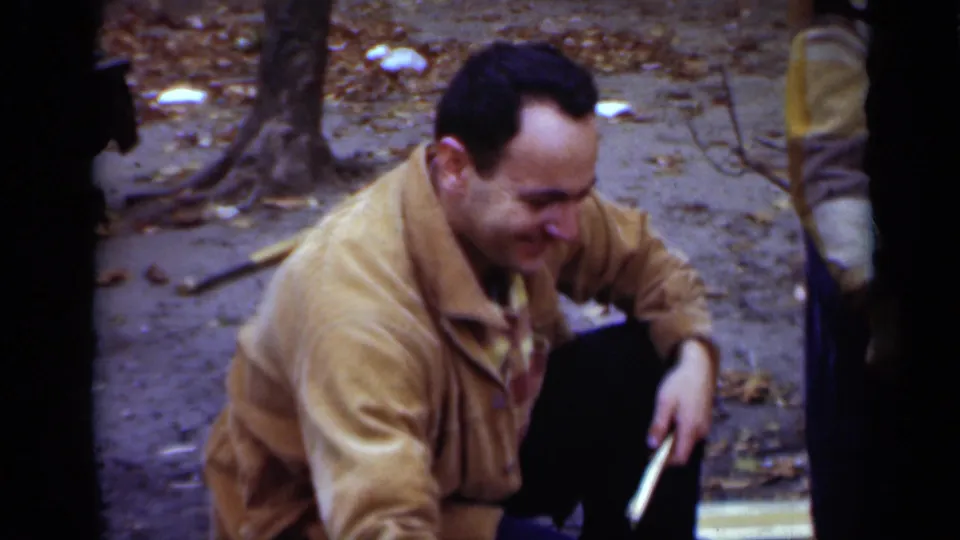 The man in a tan jacket crouches in the wooded New York setting, looking down as he handles a roasting stick near the fire area.
