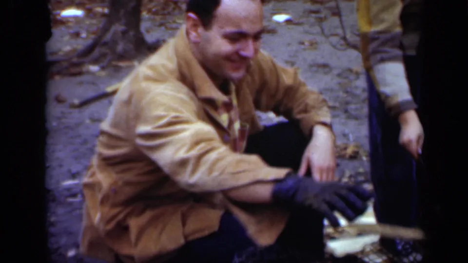 A man in a tan jacket crouches low in the wooded scene, reaching forward with a gloved hand while tending the roasting setup.