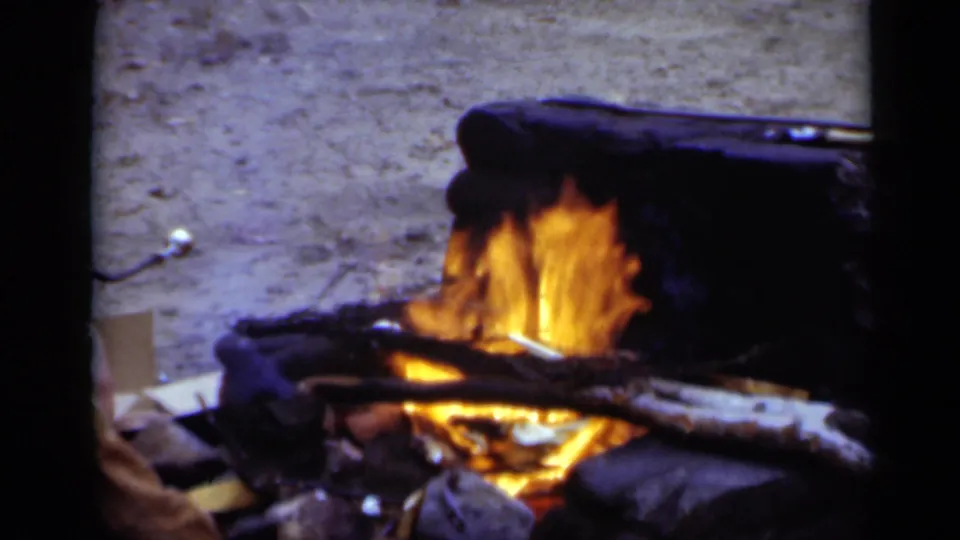 A close view shows the campfire burning in the wooded setting, with orange flames, charred wood, and a dark grate above it.