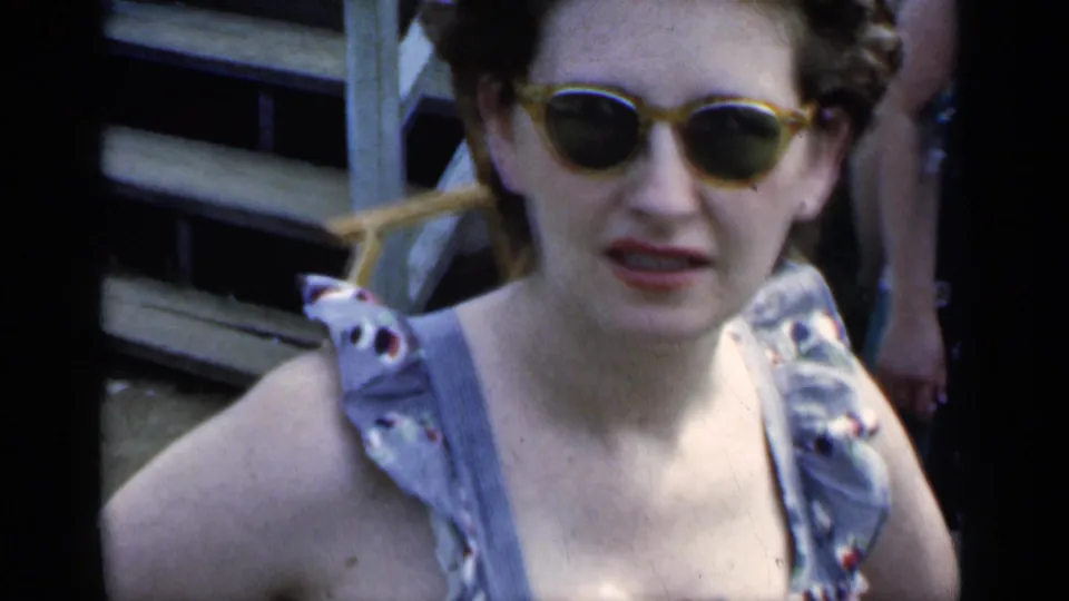 Close 1951 New York frame of a woman in yellow-framed sunglasses and a sleeveless sundress facing the camera outdoors.