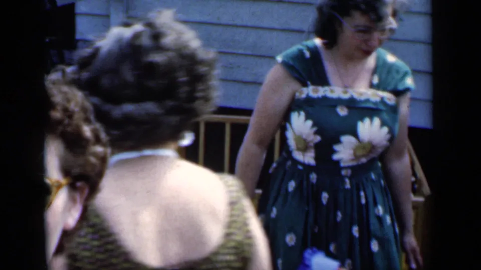 1951 New York backyard frame with two women in sleeveless dresses near house siding, wooden chairs, and a playpen.