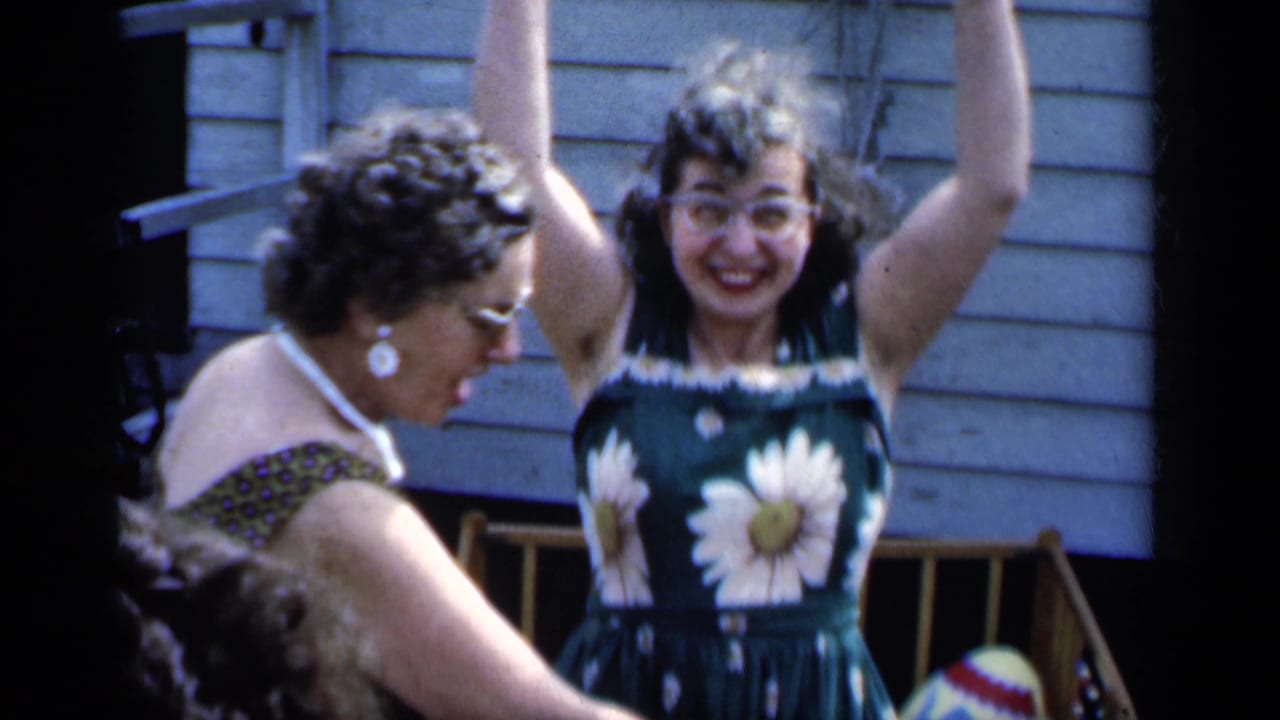 Two women in summer dresses laugh and gesture in front of a clapboard house.