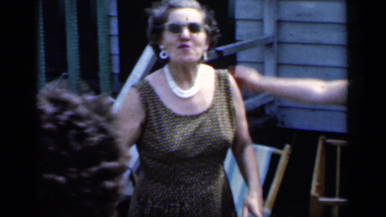 Woman in sunglasses and a patterned dress gestures beside a house and lawn chair.