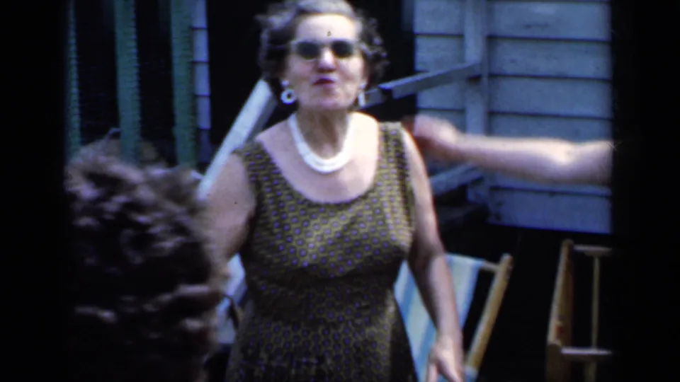 An adult woman in sunglasses and a sleeveless patterned dress gestures toward the camera in a New York backyard setting.