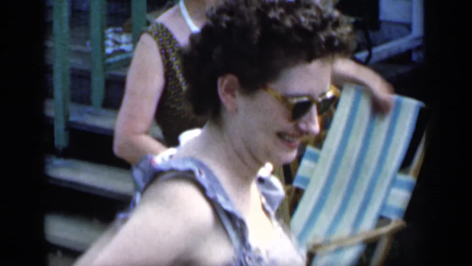 Two adult women move through the frame near a striped lawn chair, with one smiling close to the camera.