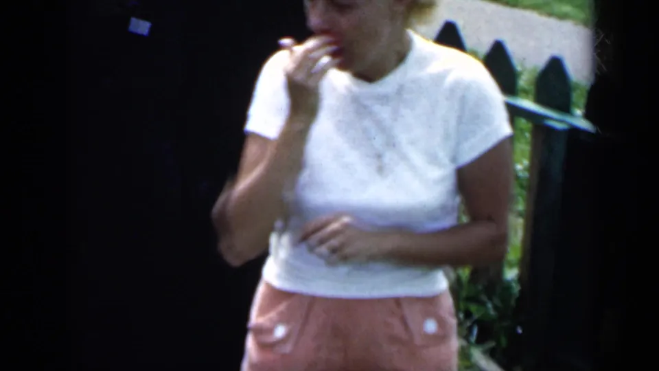 A woman in a white shirt and pink shorts laughs near a green picket fence in 1951 New York home-movie footage.