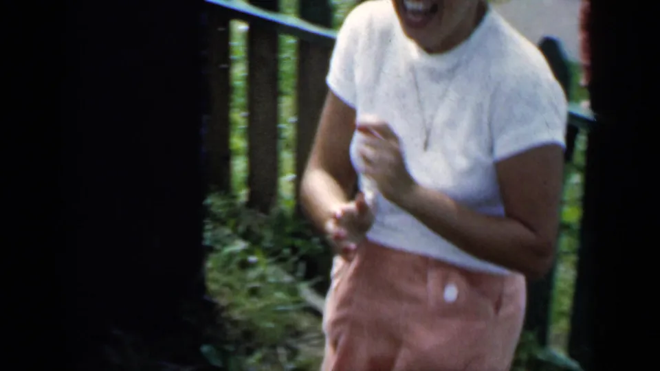 The woman continues laughing toward the camera near a green picket fence in the supported 1951 New York outdoor home-movie scene.