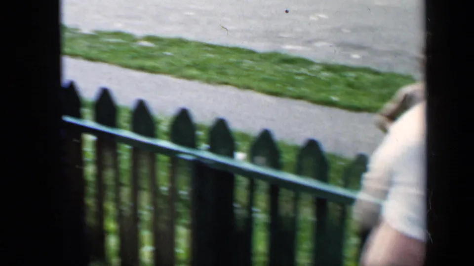 1951 New York home-movie frame with a woman partly passing a green picket fence beside grass and sidewalk.