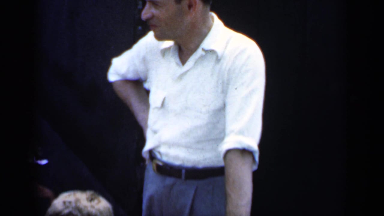 1951 New York home-movie frame of a man in a white short-sleeved shirt standing casually against a dark background.
